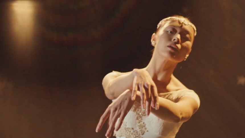 A graceful Asian ballerina in a white tutu performs a slow, elegant dance under a warm spotlight on a dark stage