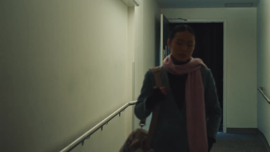 A stylish young Asian woman walks down a ramp in a dimly lit hallway towards the camera