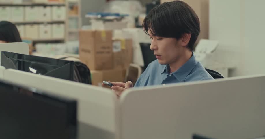 A young Asian man in a blue shirt uses his smartphone while sitting at his desk in a busy office