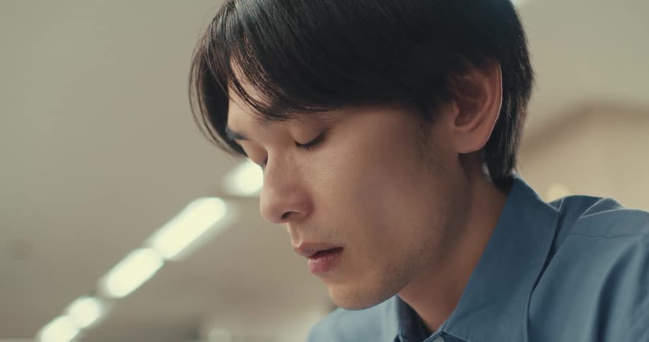 An intimate close-up shows a young Asian man with a pensive and concentrated expression
