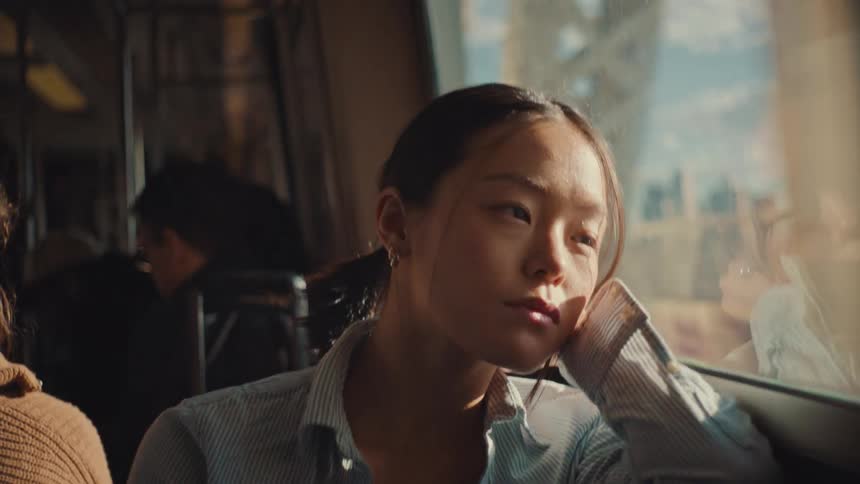 Sunlight flickers across a pensive young woman's face as she gazes out the window of a moving subway train