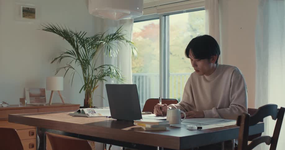 A young Asian man works from home at his sunlit dining table, using a laptop and taking notes