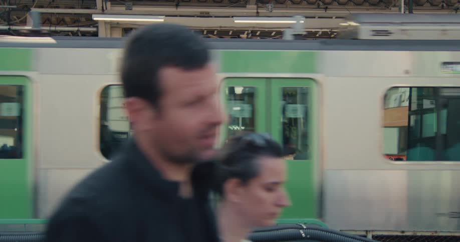 Side view of a silver and green train speeding past a busy station platform in Japan as commuters walk by