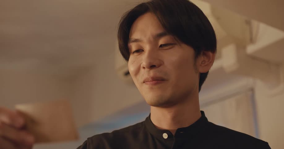 A young East Asian man's expression softens into a fond smile as he looks at a nostalgic memento