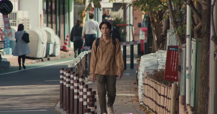 A young Japanese man walks in slow motion down a sunlit city sidewalk with other pedestrians in the background