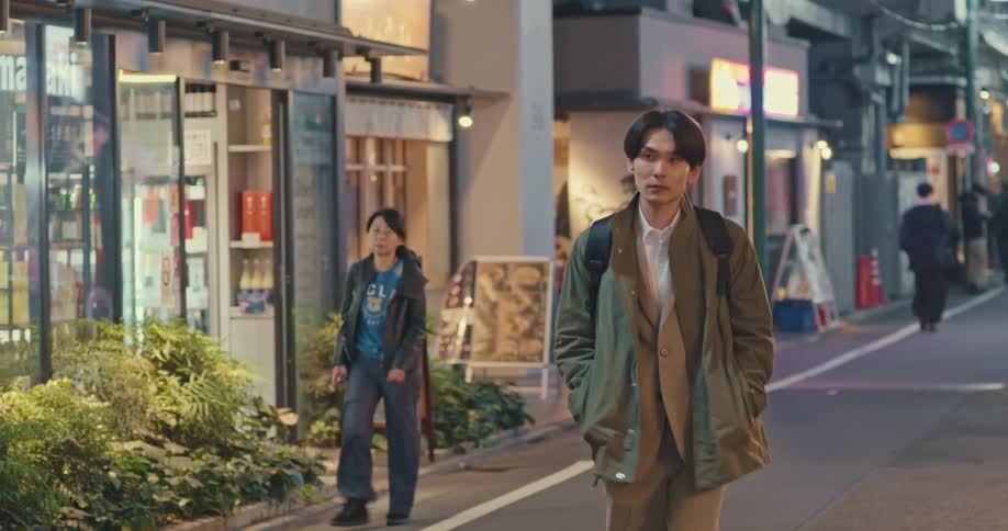A stylish young Asian man walks in slow motion down a quiet, illuminated alleyway lined with shops in a Japanese city at night