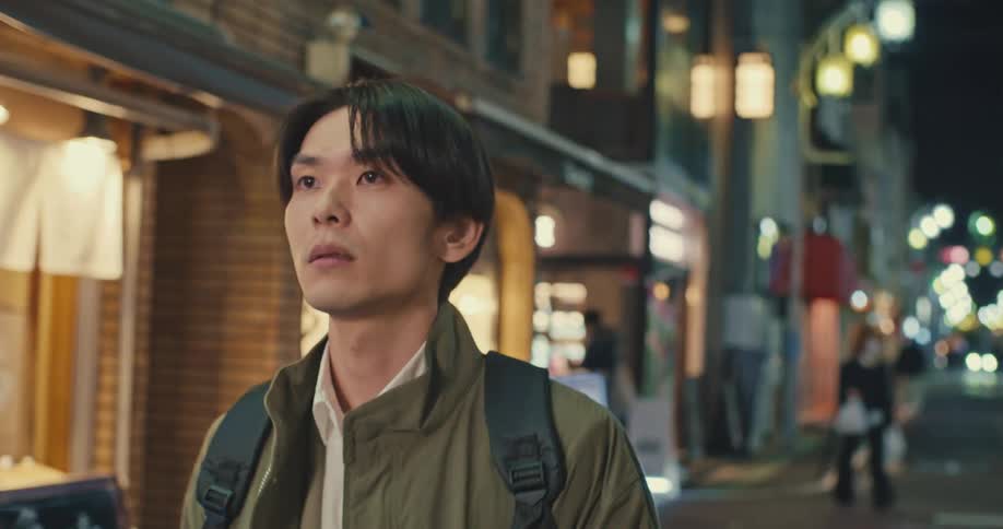 A pensive young Japanese man with a backpack walks slowly down an illuminated city street at night