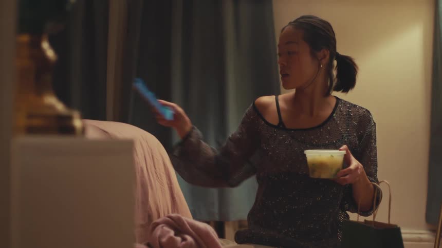 A young Asian woman kneels on her bedroom floor, pulls takeout food from a bag, and takes a happy selfie with it