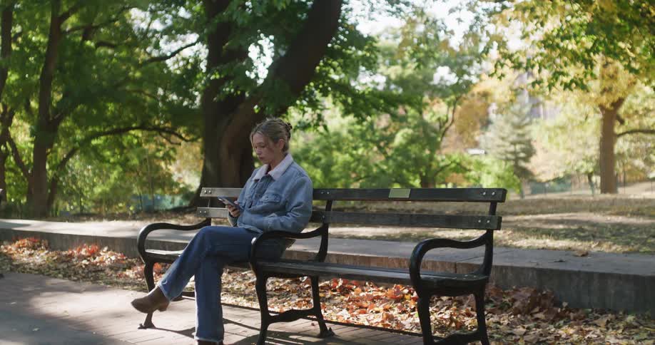 A young woman sits on a park bench in the autumn sunlight, looking down at her smartphone