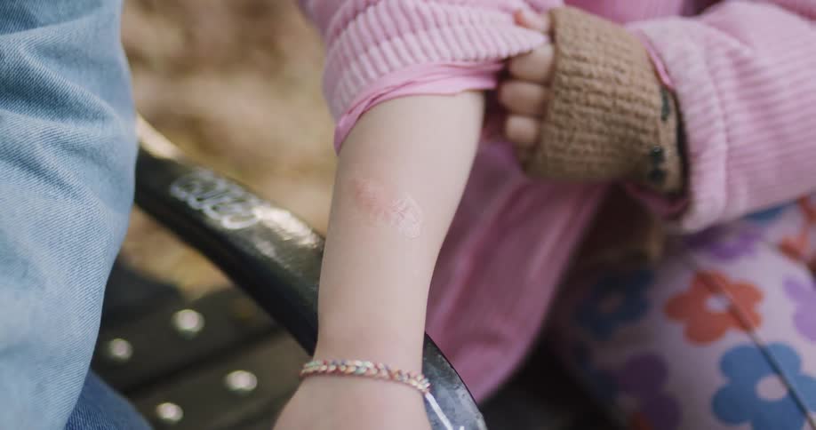 Close-up of a child in a pink jacket pulling up their sleeve to show a heart-shaped bandage on their arm