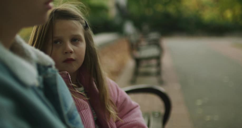 A sad little girl looks on as her mother is distracted by her phone while they sit together on a park bench