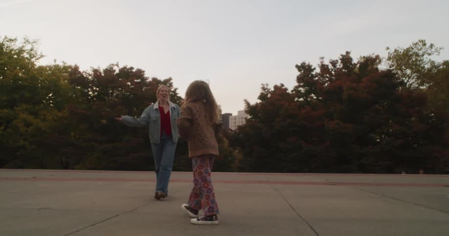 A joyful young girl performs a cartwheel in slow motion for her clapping mother in a city park at dusk