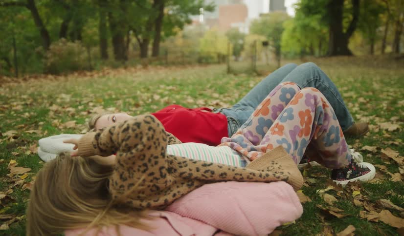 A mother and her young daughter share a quiet, loving moment as they lie together on the grass amongst fallen autumn leaves in a park