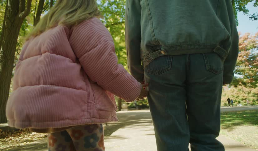 A young girl reaches for her mother's hand as they walk together through a sunlit park path in autumn