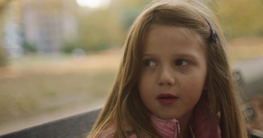 A close-up slow-motion shot of a sad little girl with a tear on her cheek sitting on a park bench