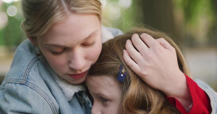 A compassionate young mother tenderly comforts her sad daughter, pulling her into a warm and reassuring hug while sitting in a park