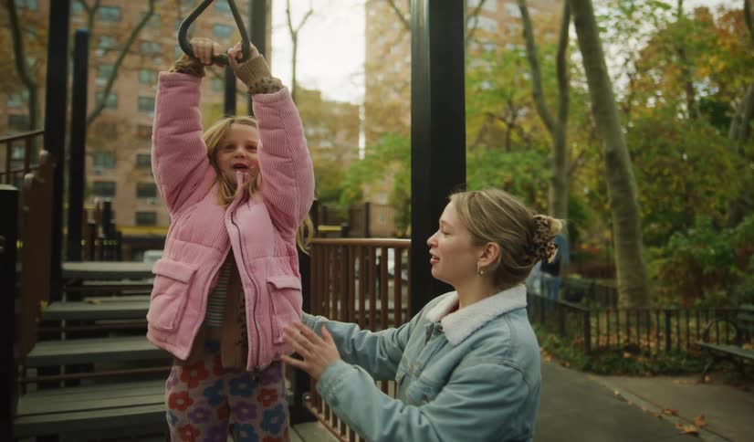 A smiling mother encourages her young daughter as she playfully hangs from a monkey bar ring at a city playground in slow motion