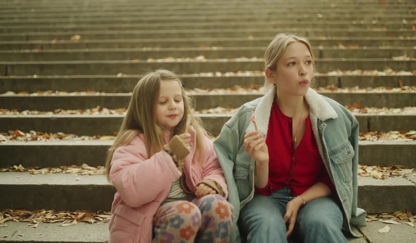 A young woman and a little girl happily eat lollipops together while sitting on concrete steps covered in autumn leaves in slow motion