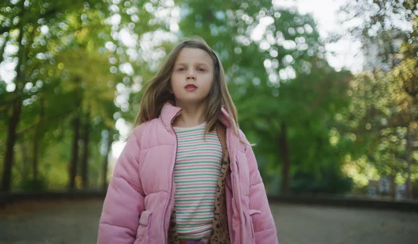 A confident young girl in a stylish pink jacket walks towards the camera in slow motion through a park covered in autumn leaves