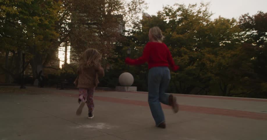 A carefree mother and her young daughter playfully run and jump in the air in slow motion at a park