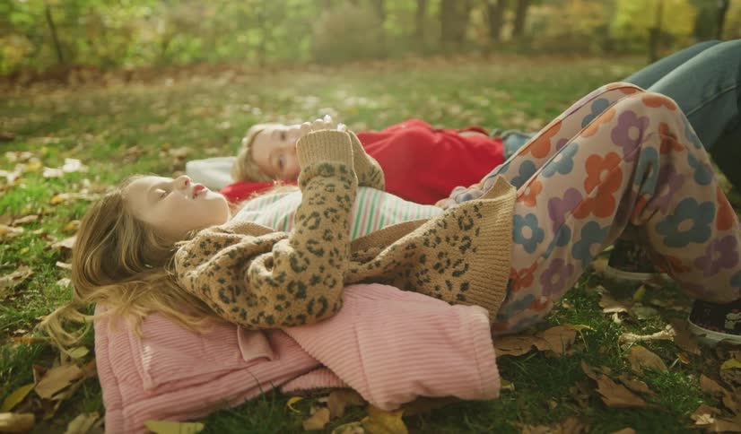 Two young sisters share a peaceful moment as they lie together on the grass in a sunlit park during autumn