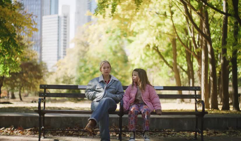 A mother and her young daughter have a serious conversation while sitting together on a park bench on a sunny autumn day in the city
