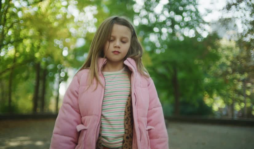 A stylish young girl in a pink corduroy jacket walks confidently down a leaf-strewn path in a park on a sunny autumn day