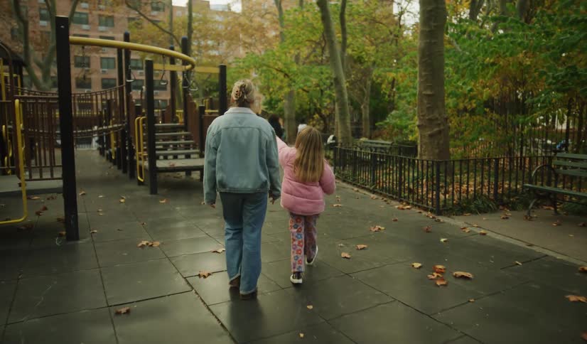 A mother and her young daughter walk together through a city playground on a cool autumn day