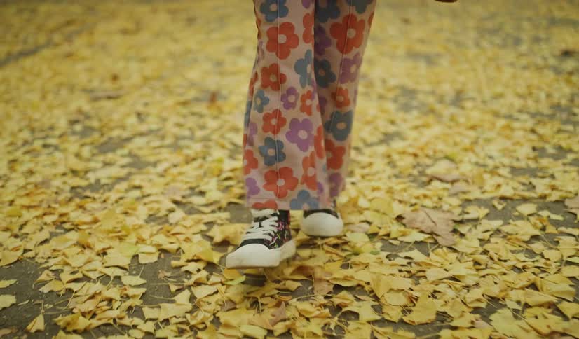 A little girl in colorful floral pants walks through a path covered in yellow leaves and kneels to play with them in an autumn park