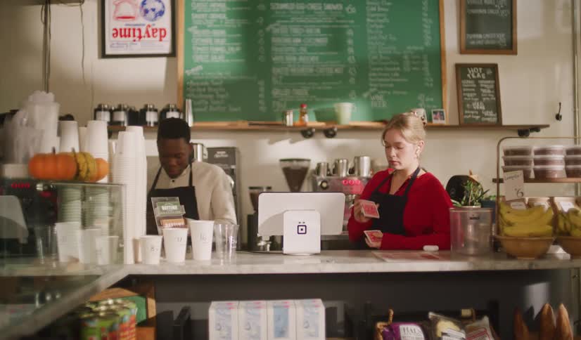 A female barista shuffles a deck of cards at a coffee shop counter while her male coworker approaches and talks to her
