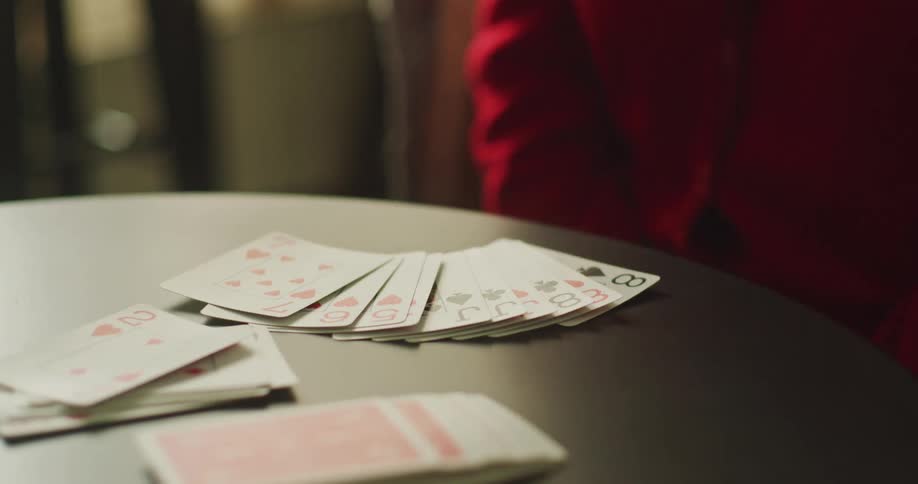 A close-up of a card player in a red sweater fanning out their hand onto a dark table before gathering the cards back up