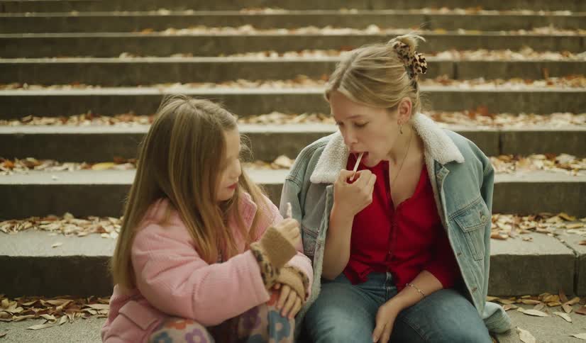A young girl and her mother share a sweet moment eating candy while sitting on leaf-covered steps in a park during autumn