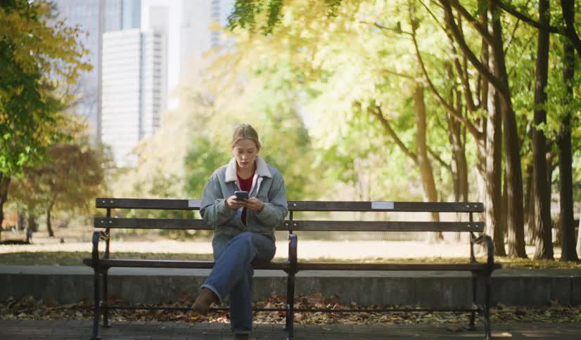 A young woman walks to a park bench on a sunny autumn day in the city, sits down, and waits