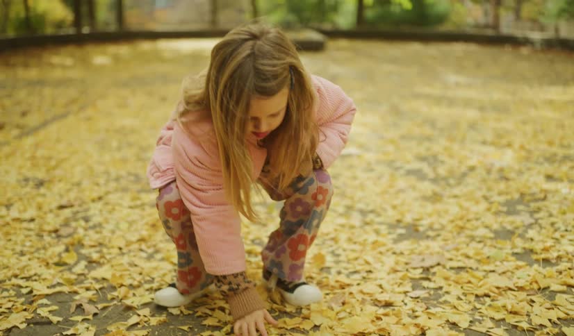 A young girl in a pink jacket and floral pants walks through a park covered in yellow autumn leaves, stopping to pick one up