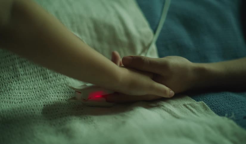 A supportive person gently holds the hand of a patient wearing a glowing pulse oximeter while resting in bed