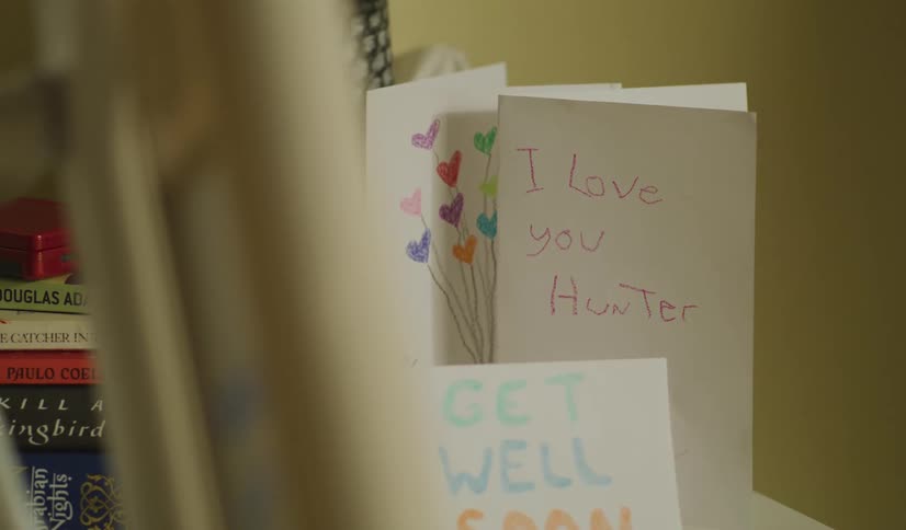 Handmade "Get Well Soon" and "I Love You" cards from a child sit on a nightstand next to a stack of books