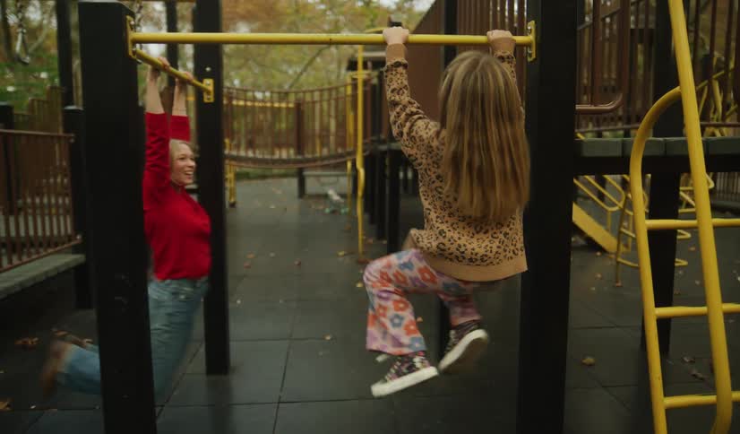 A playful mother and her young daughter laugh and swing from the monkey bars together at a park playground on a cool autumn day
