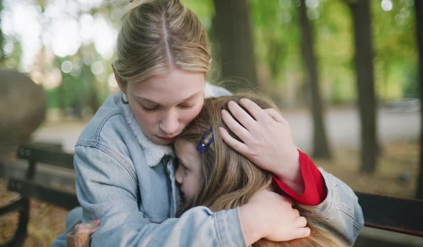 A caring young woman embraces and comforts a sad little girl while sitting on a park bench