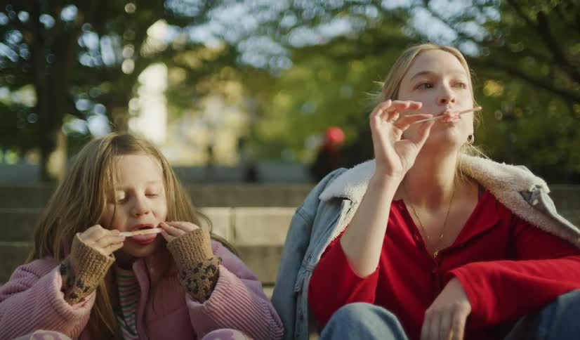 A young girl and her mother sit together on park steps in slow motion, playfully enjoying an ice cream treat and making silly faces with the popsicle sticks
