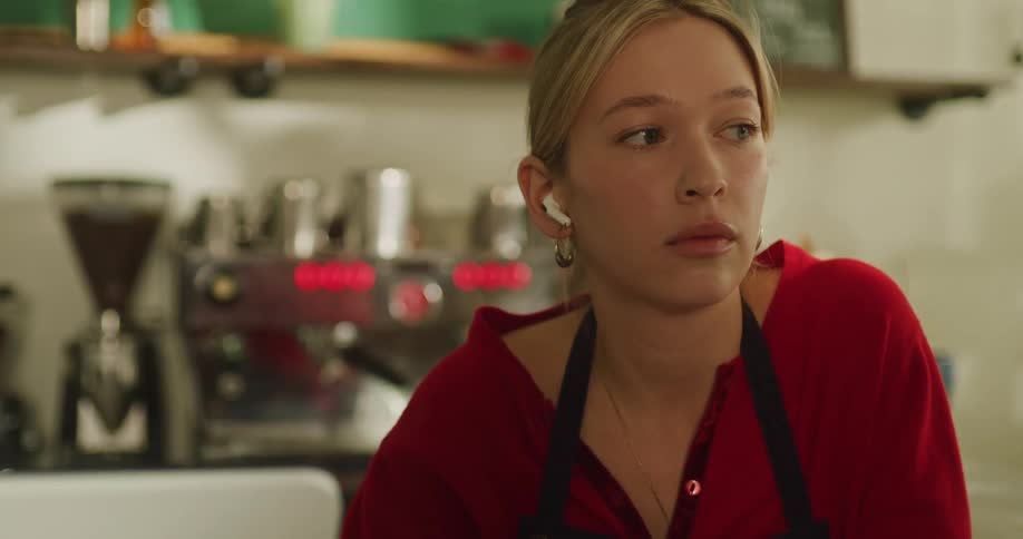 A bored young barista with wireless earbuds idly shuffles cards at the counter of a quiet coffee shop