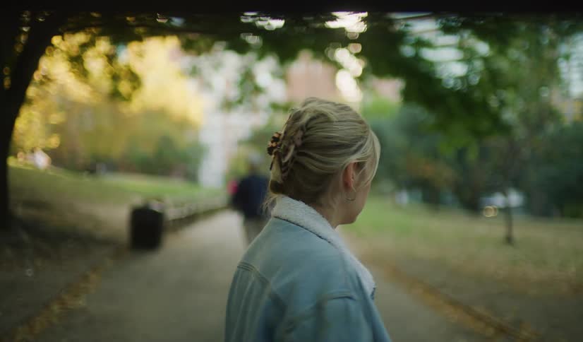 A pensive young woman walks through a sunlit city park, her worried expression turning to one of alarm as she stops and looks around