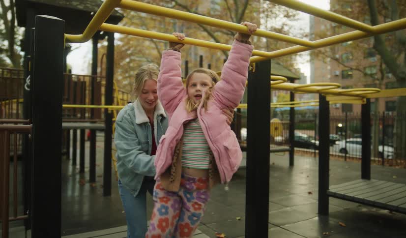 A mother supports her young daughter as she joyfully jumps up to hang from the monkey bars at a city park playground in slow motion