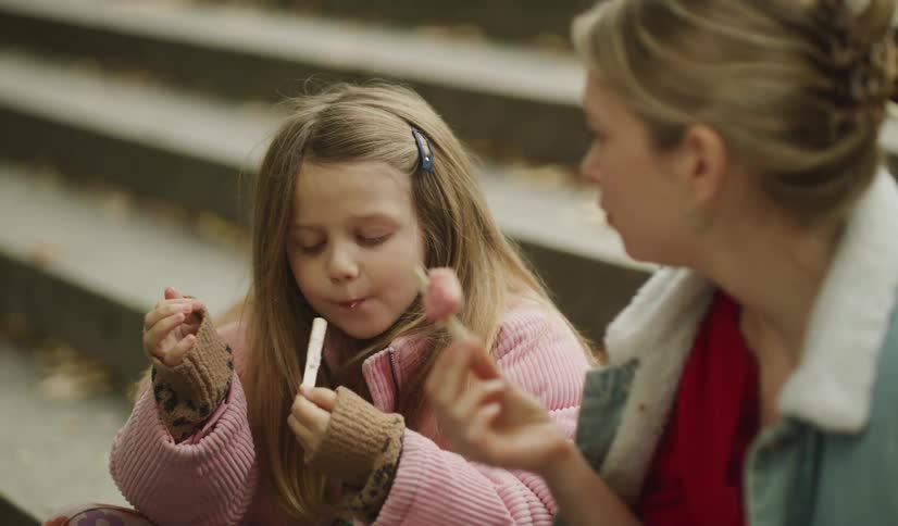 A young girl with long hair enjoys a sweet pink treat on a stick with her mother while sitting on outdoor steps in slow motion