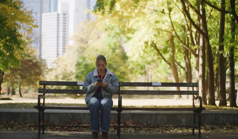 A young woman takes a seat on a park bench to relax, pulling out her phone to scroll amidst the autumn foliage of a city park