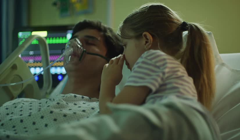 A worried young daughter sits in bed and prays over her unconscious father who is on life support in the hospital