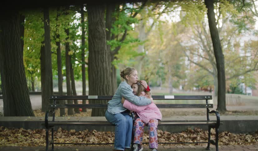 An older sister consoles her upset younger sister with a comforting hug while sitting together on a park bench in autumn