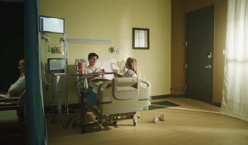 A young girl visits her older brother in his hospital room while their mother sits nearby