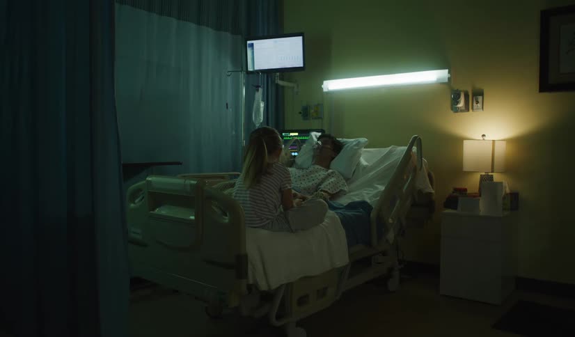 A caring young daughter visits her sick mother in a dimly lit hospital room at night