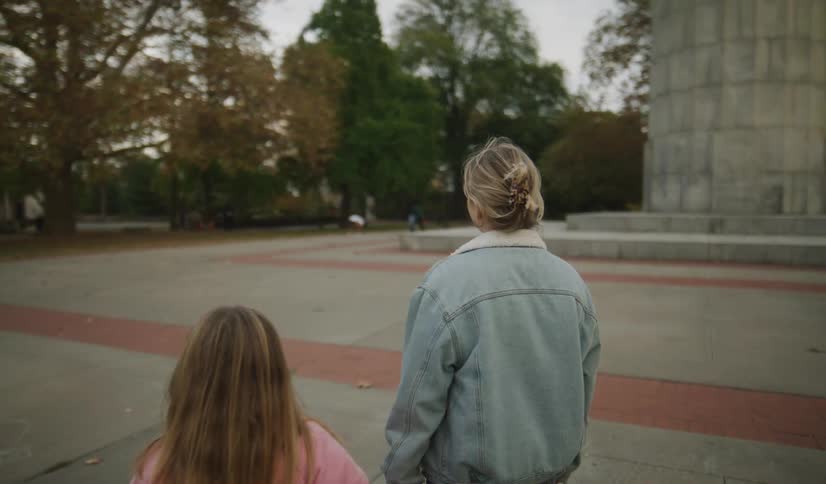 A mother and daughter walk hand-in-hand through an autumn city park, before the camera focuses on the thoughtful young woman standing alone