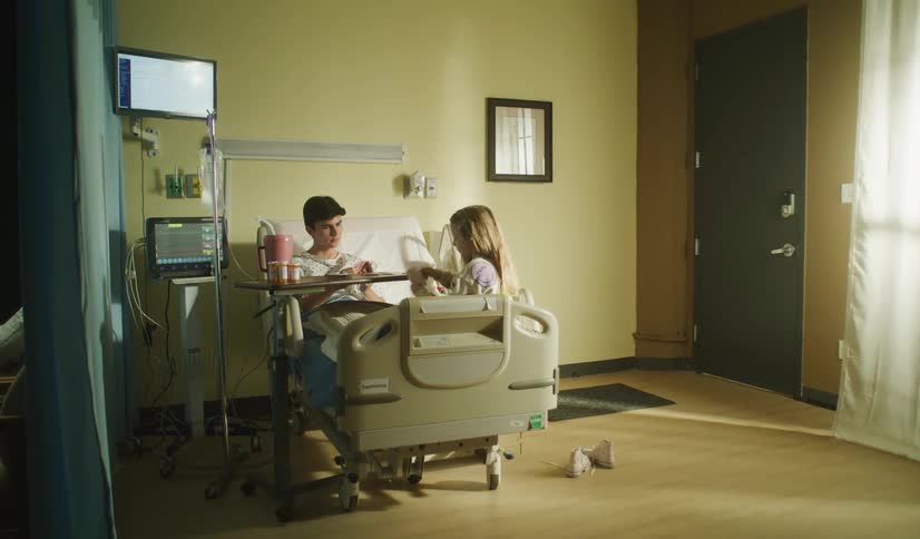 A young girl visits her older brother in a hospital room, keeping him company while he recovers as their mother sits nearby